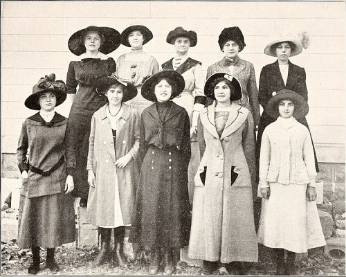 A group of ten women in early 20th-century attire, wearing hats and coats, posing outdoors.