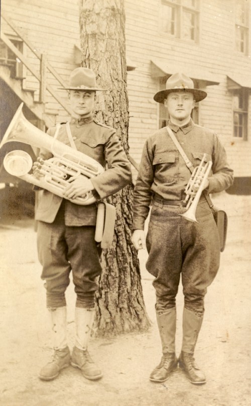 Two soldiers in vintage military uniforms stand beside a tree, one holding a tuba and the other a trumpet.