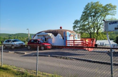 A quirky ice cream shop shaped like a sundae, with cars parked outside and a scenic view in the background.