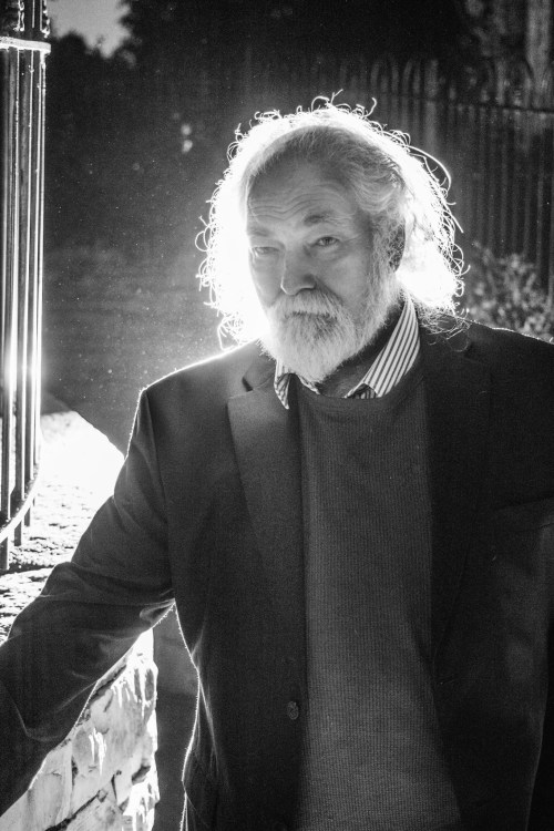A bearded man in a suit stands by a stone wall, illuminated from behind, creating a dramatic silhouette effect.
