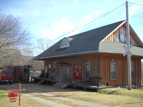 A rustic wooden building with a sloped roof, red doors, and a nearby sign, set in a rural landscape.