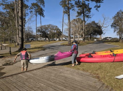 Two people launching kayaks at a park, surrounded by trees and a campground in the background.