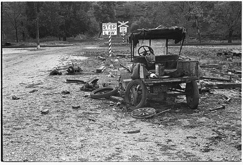 A damaged, abandoned car sits near a railroad crossing sign, surrounded by debris and a desolate landscape.