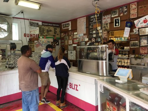 A small ice cream shop with customers at the counter, a server preparing orders, and colorful decor on the walls.