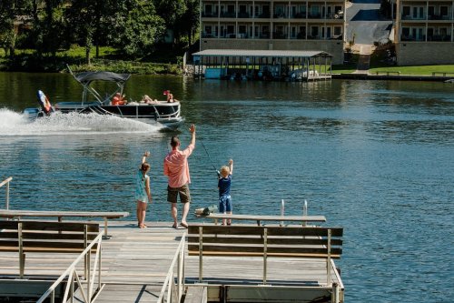 A family waves from a dock as a boat speeds by on a sunny lake day.