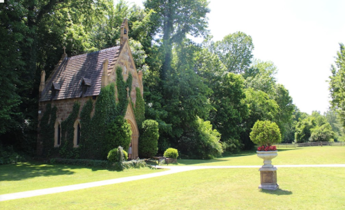 A quaint stone chapel covered in ivy, surrounded by lush greenery and a decorative planter in a sunny park setting.