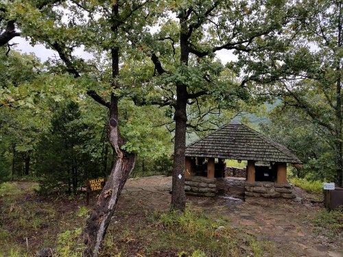 A rustic shelter with a shingled roof surrounded by trees on a misty day, with a trail sign nearby.