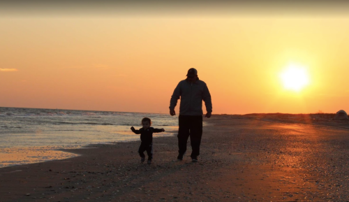 A silhouette of an adult and a child walking on a beach at sunset, with waves gently lapping at the shore.