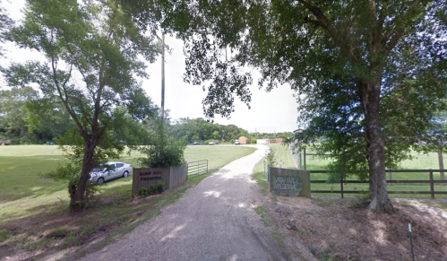 A gravel driveway leads to a green field, framed by trees and a gate labeled "Preserve." A car is parked nearby.