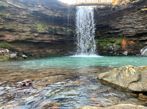 A serene waterfall cascades into a turquoise pool, surrounded by rocky terrain and lush greenery.