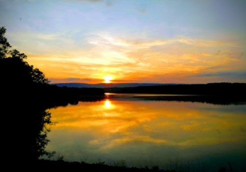 A serene sunset over a calm lake, reflecting vibrant colors in the water, surrounded by trees and distant hills.
