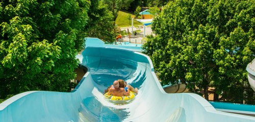 Two people slide down a blue water slide surrounded by lush green trees, with a water park visible in the background.