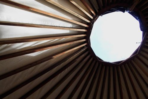 View from inside a yurt, showing wooden beams and an octagonal opening at the top with a clear sky visible.