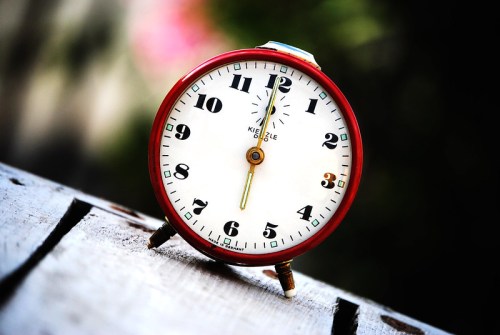 A vintage red alarm clock with a white face, black numbers, and gold hands, set against a blurred background.