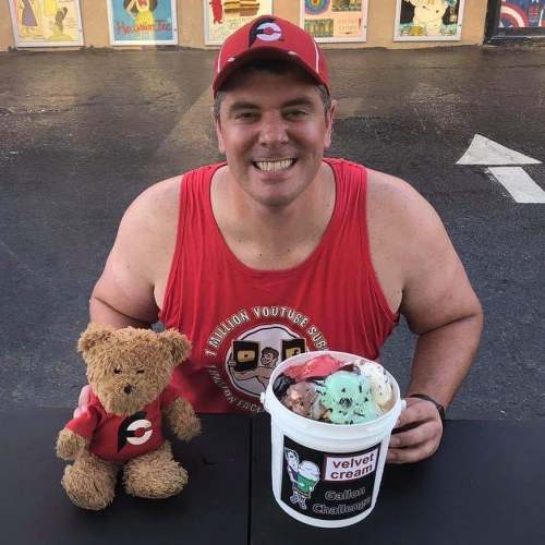 A smiling man in a red tank top and cap sits at a table with a teddy bear and a bucket of ice cream.