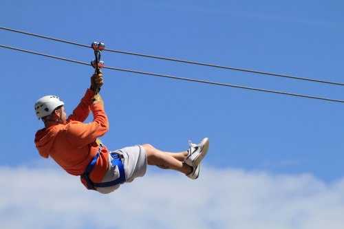 A person in an orange hoodie and helmet zip-lining against a clear blue sky.