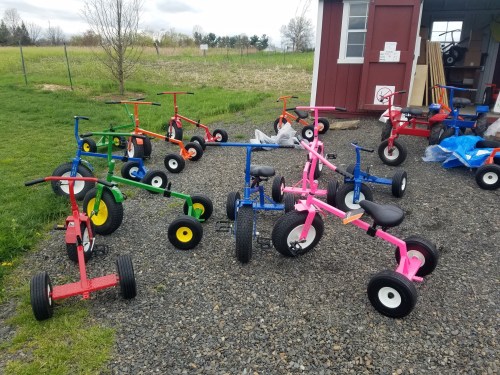 A variety of colorful tricycles parked on gravel outside a shed, with green fields in the background.
