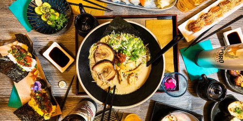 A vibrant spread of Japanese dishes, featuring ramen, sushi, and various sides, with drinks and garnishes on a wooden table.
