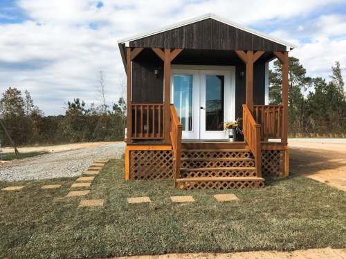 A small, dark wooden cabin with a porch and steps, surrounded by grass and gravel, under a cloudy sky.