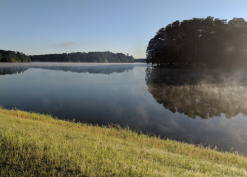 A serene lake at dawn, with mist rising from the water and trees lining the shore under a clear blue sky.