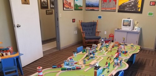A colorful playroom with a train set, toy buildings, and a rocking chair, featuring framed artwork on the walls.