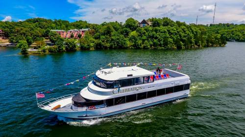 A large boat with colorful flags cruising on a lake, surrounded by green trees and a blue sky.