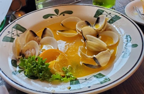 A bowl of clams in a yellow broth, garnished with lettuce and orange slices, served on a decorative plate.