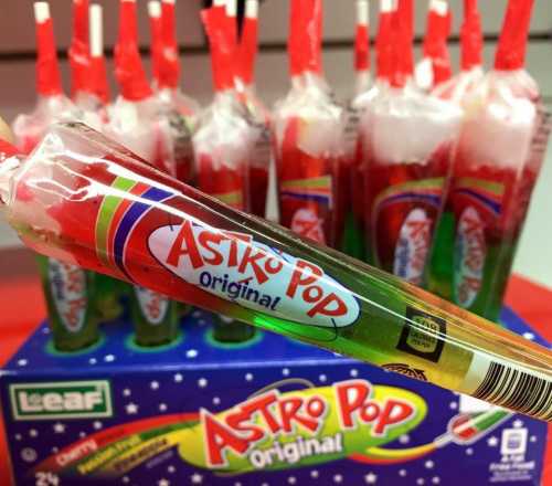 A close-up of an Astro Pop candy with a colorful wrapper, in front of a box containing more Astro Pops.