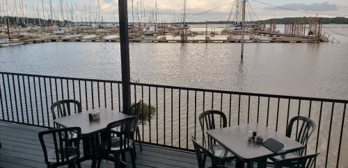 View of a marina with boats docked, seen from a deck with empty tables and chairs overlooking the water.