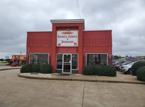 A red building with a sign reading "Kountry Bakery Restaurant," surrounded by greenery and parked cars.