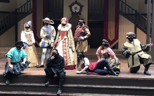 A group of performers in historical costumes poses on stage, all wearing masks, in a theatrical setting.