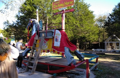 A large, colorful rocking horse in a park, with a ladder and people nearby, surrounded by trees and festive decorations.