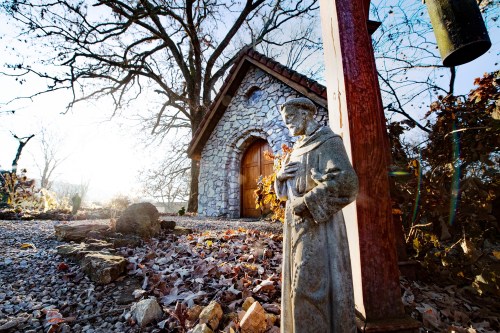 A stone chapel surrounded by trees, with a statue of a saint in the foreground, illuminated by soft sunlight.
