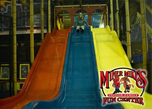 A person sits at the top of colorful slides in an indoor play center, ready to slide down. Miner's Mike's logo is visible.