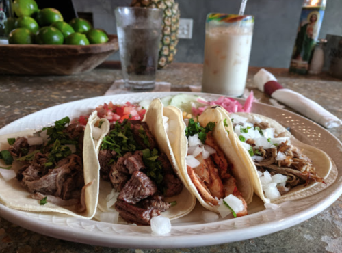 A plate of four tacos filled with various meats, topped with onions and cilantro, alongside a drink and a glass of water.
