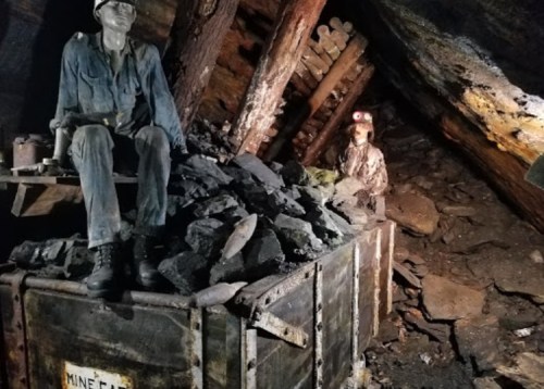 A dimly lit mine interior featuring two figures, one seated on a cart filled with coal, surrounded by wooden beams.