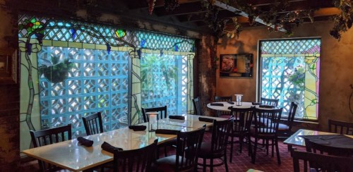 Cozy restaurant interior with stained glass windows, tables set for dining, and greenery visible outside.