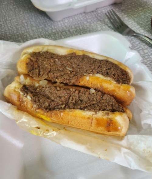 Two hot dog buns filled with a thick, brown meat mixture, garnished with chopped onions, on a white takeout container.