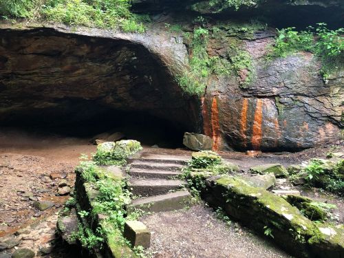 A rocky cave entrance surrounded by greenery, with stone steps leading into the cave and orange streaks on the rock face.