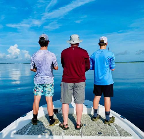 Three people fishing from a boat, facing the calm water under a blue sky with scattered clouds.
