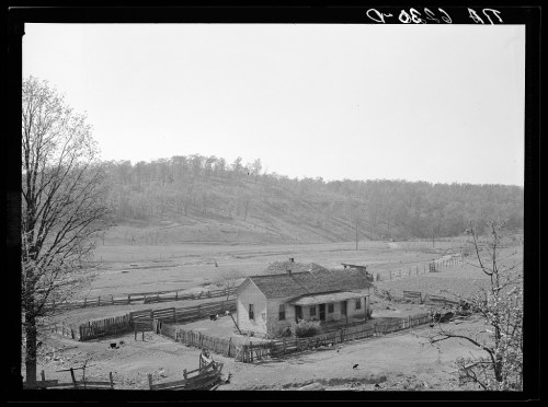 A rural farmhouse surrounded by a wooden fence, with hills and trees in the background, captured in black and white.