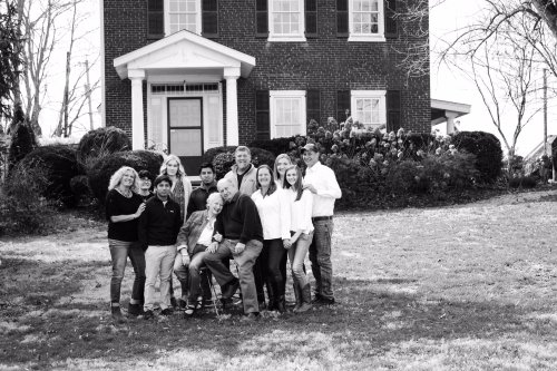 A group of people poses together in front of a large brick house, surrounded by greenery, in a black and white photo.