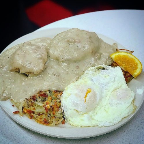 A plate of biscuits and gravy, hash browns, and a fried egg, garnished with an orange slice.
