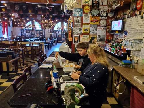 A man and a woman at a bar counter, reviewing food orders in a lively restaurant filled with vintage decor.