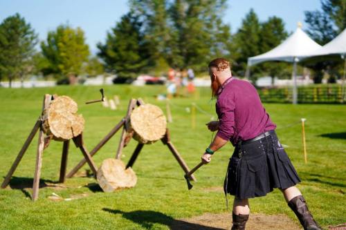 A man in a kilt throws an axe at wooden targets in a grassy field during a sunny outdoor event.
