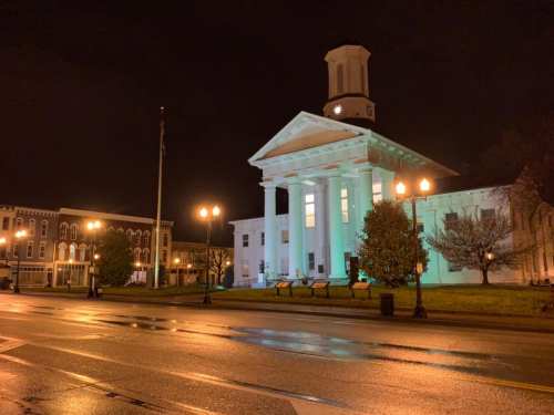 Historic building illuminated at night, featuring columns and a clock tower, with a quiet street in the foreground.
