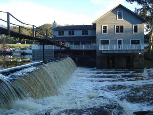 A watermill with a flowing waterfall in front, surrounded by trees and a blue sky.