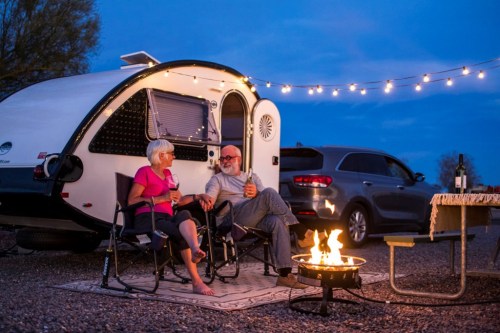 A couple sits by a campfire outside their camper, enjoying drinks and each other's company under string lights at dusk.