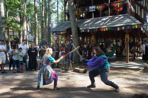 Two performers in colorful costumes engage in a playful sword fight, surrounded by an audience in a forested setting.
