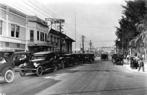Black and white photo of vintage cars lined up on a street, with buildings and power lines in the background.
